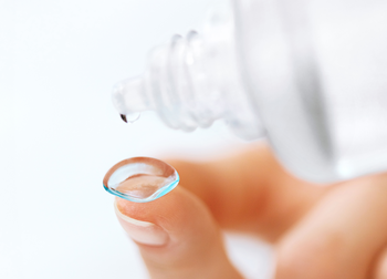 Das Bild zeigt Finger mit einer Linse und eine Flasche Linsenpflegemittel.
