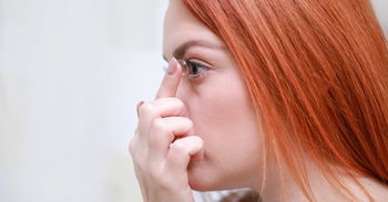 Zu sehen ist eine rothaarige Frau, die sich eine Kontaktlinse ins Auge einsetzt