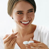 Das Bild zeigt eine junge lachende Frau mit einem Kontaktlinsenbehälter und einer Kontaktlinse in der Hand.