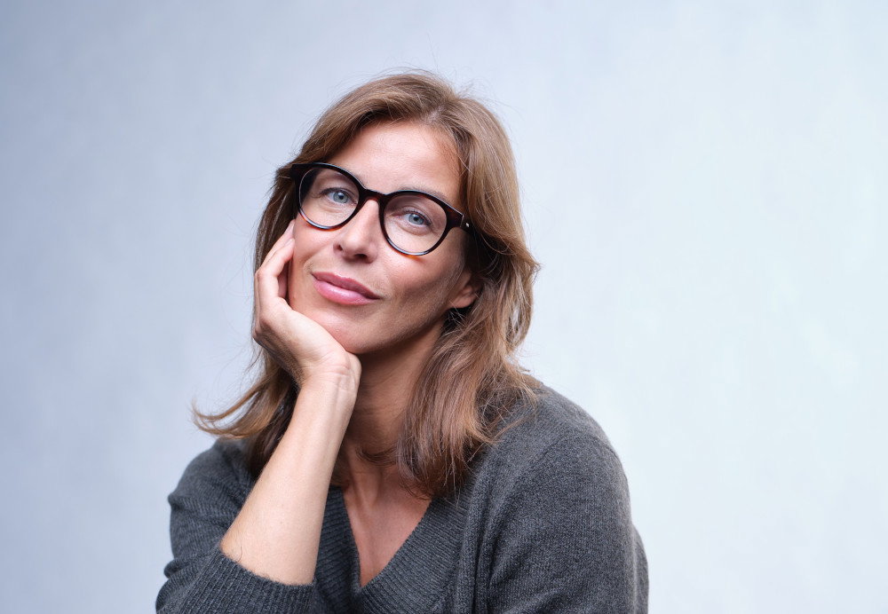 Eine Frau mit Brille und grauem Pullover stützt ihr Kinn auf ihrer Hand ab.
