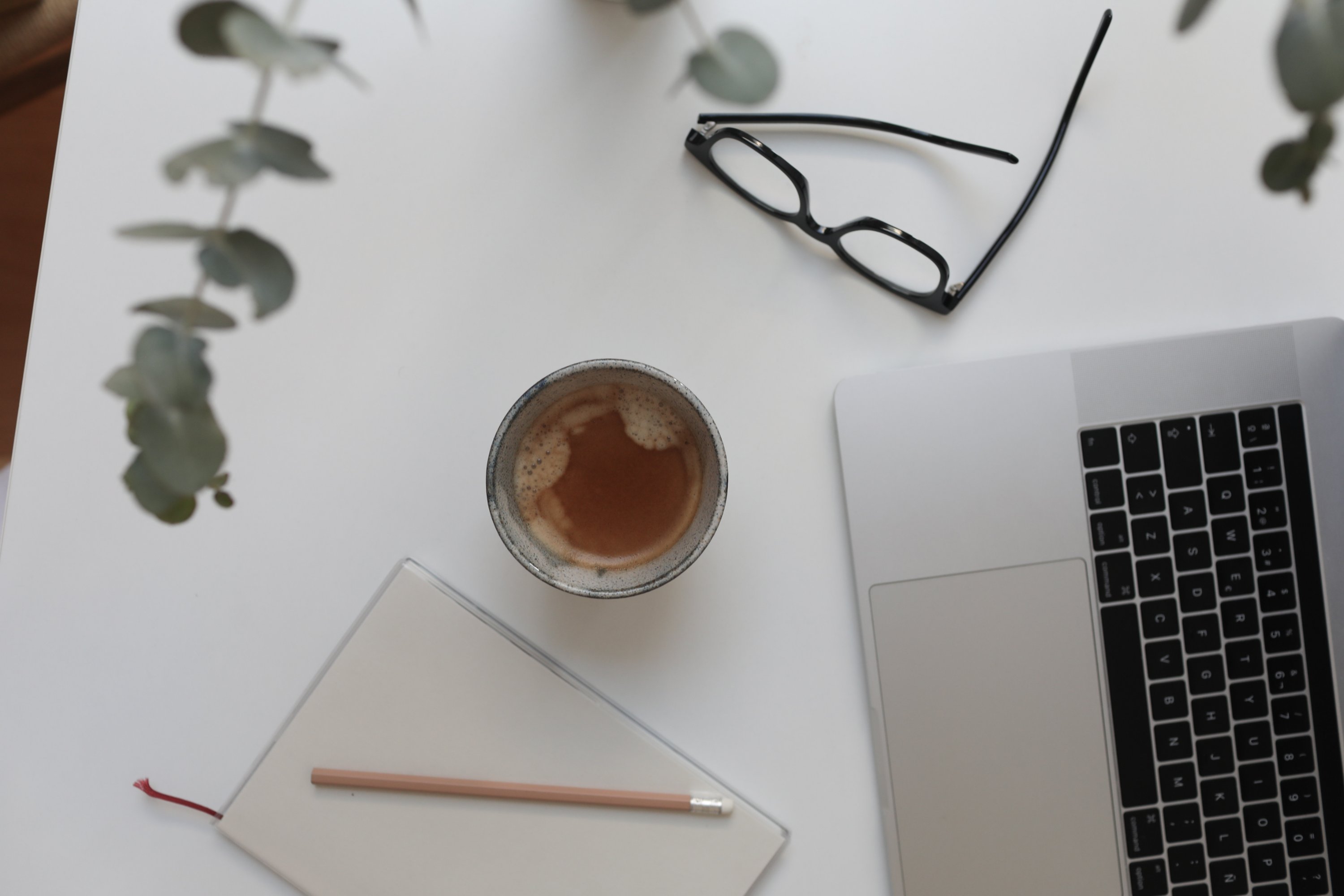 Im Bild ist eine schwarze Brille, ein Laptop, ein Blog mit Stift und eine Tasse zu sehen