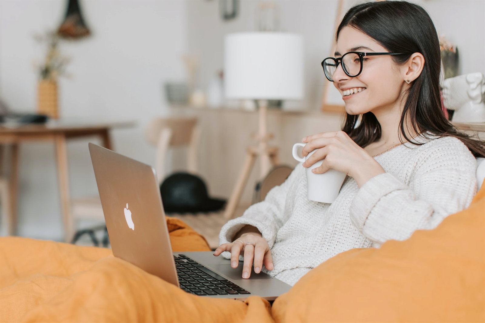 Eine Frau mit Tasse und Brille, die auf dem Sofa in ihren Laptop schaut.